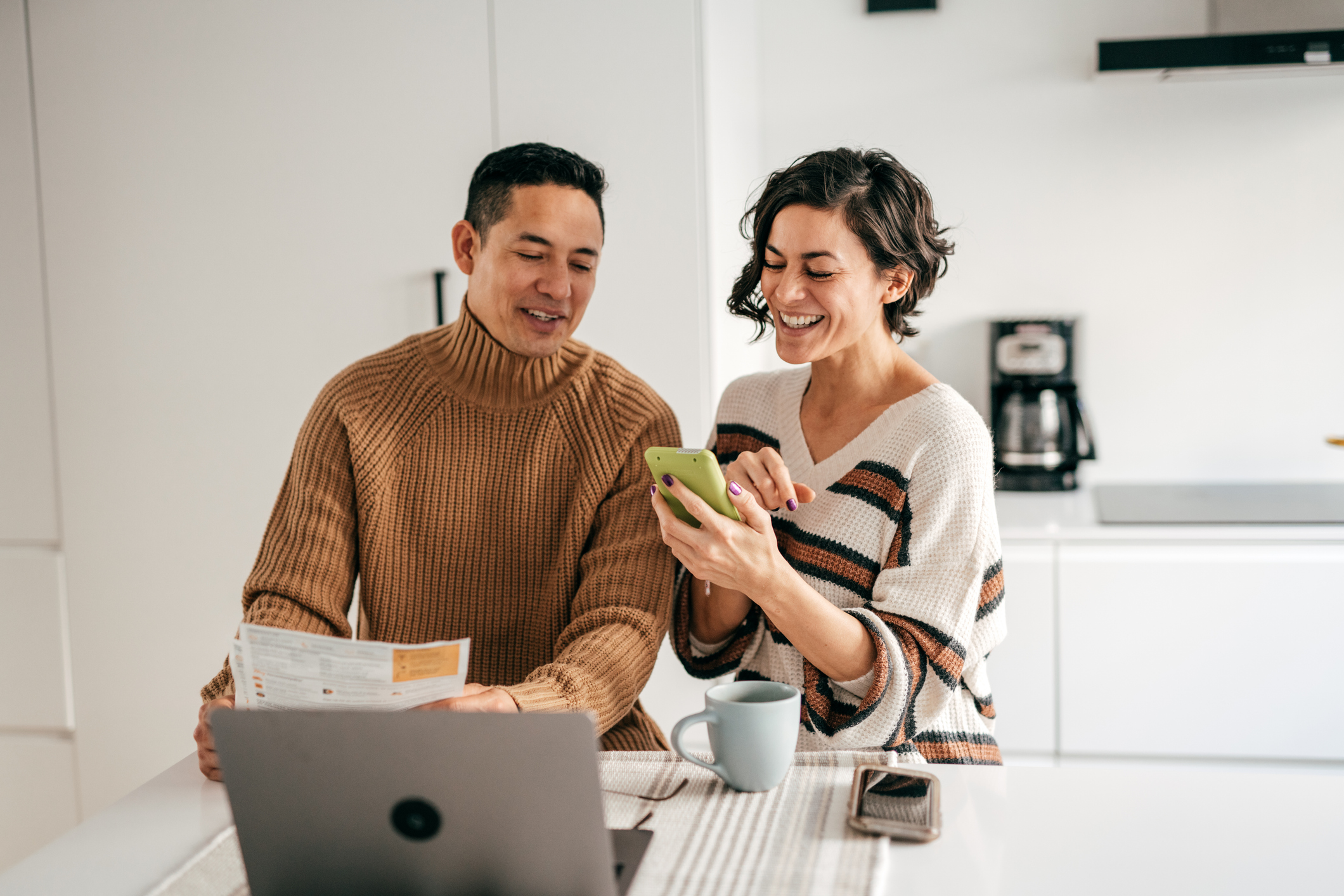 A couple sitting at a kitchen table with a laptop and paperwork, while the woman shows something on her phone to the man
